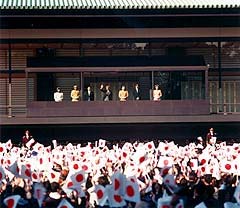 Seven members of the Imperial Family stand on a balcony while hundreds below waive Japanese flags