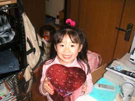 A young girl, standing in a small room, looks up, smiling, while holding a heart shaped box