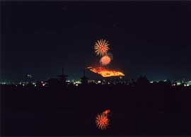Photo at night of fireworks exploding over a large burning area of a mountain