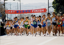 Hundreds of runners, wearing sashes and numbers leaving the starting gate of an ekiden