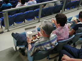 A couple, sitting at a stadium, eat sushi from a box, while watching a game