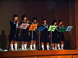 Six girls in uniform on a stage, singing