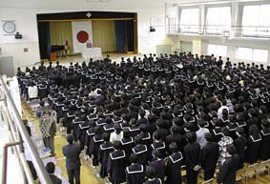 Students and teachers standing in gym, facing stage, listening to a speech