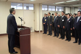 Men in suits stand, listening to a speech