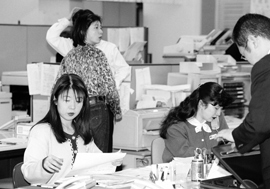 たくさんの書類と機械に囲まれたオフィスで働く女性たちの白黒写真。