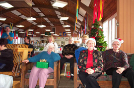 クリスマスパーティー会場に置かれた木のいすに座って微笑む、サンタの帽子をかぶった高齢の参加者たち。
