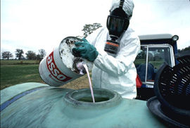 A worker wears a gas mask while pouring chemicals.