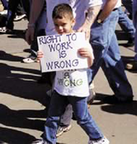 白人の少年が小さなプラカードを胸の前に持ち、その背後にはデモに参加している人たちの足が見えます。
