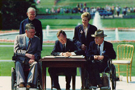 車いすにすわった２人の男性に見守られながら、ブッシュ大統領（シニア）が署名しています。